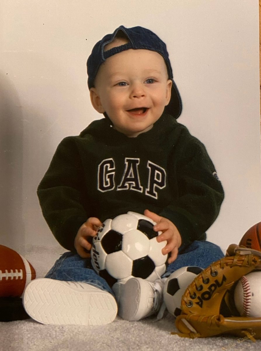 Baby Ethan in a backwards cap holding a soccer ball, surrounded by sports gear